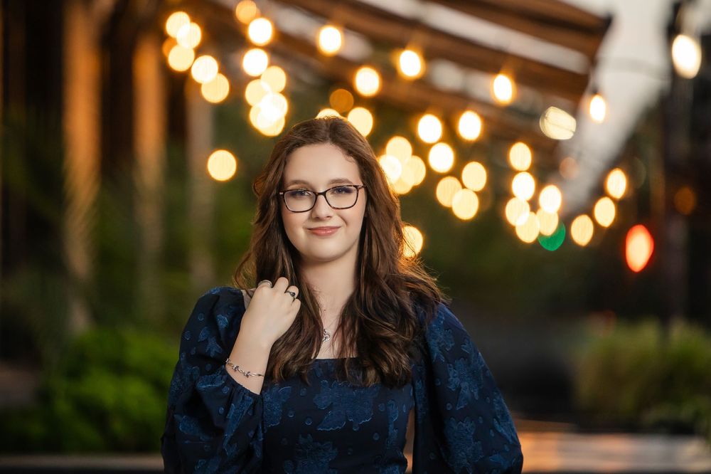 Smiling young woman in navy floral top stands under warm string lights at dusk.
