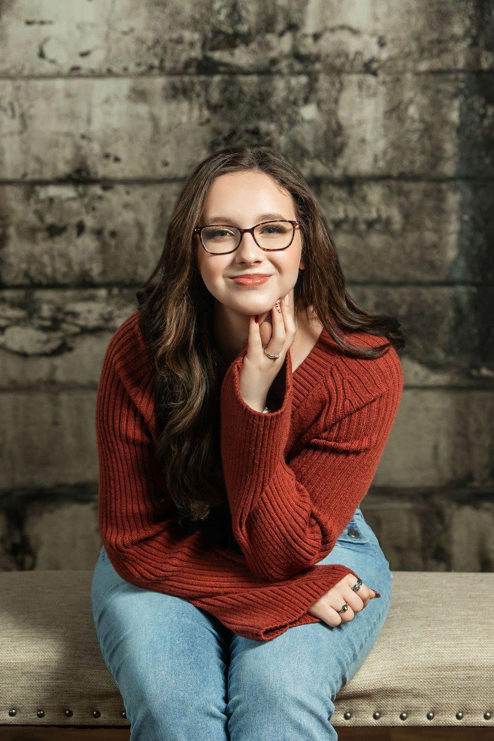 Smiling young woman in red sweater and jeans sits on bench against rustic concrete wall.