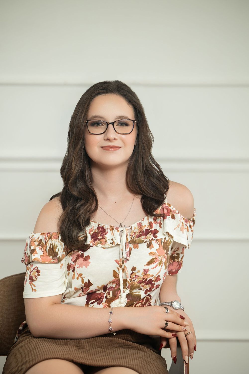 Young woman with glasses in a floral off-shoulder top seated and smiling against a white background.