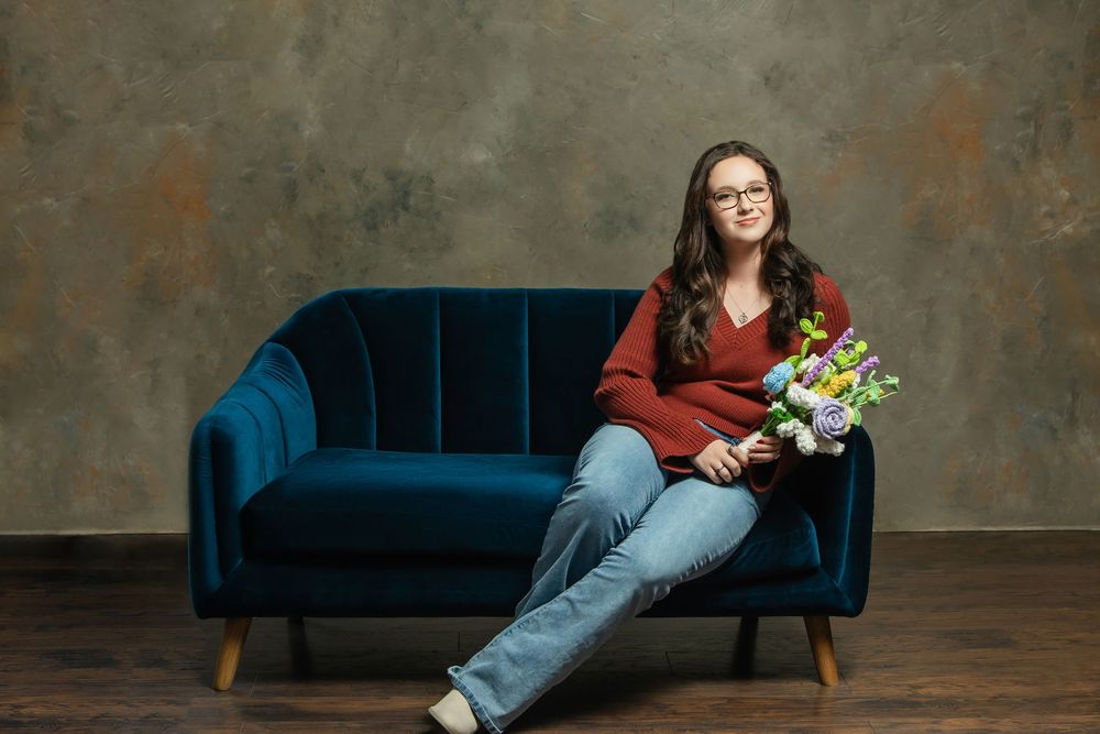 Young woman with glasses sitting on a blue velvet sofa holding colorful flowers in a studio setting.