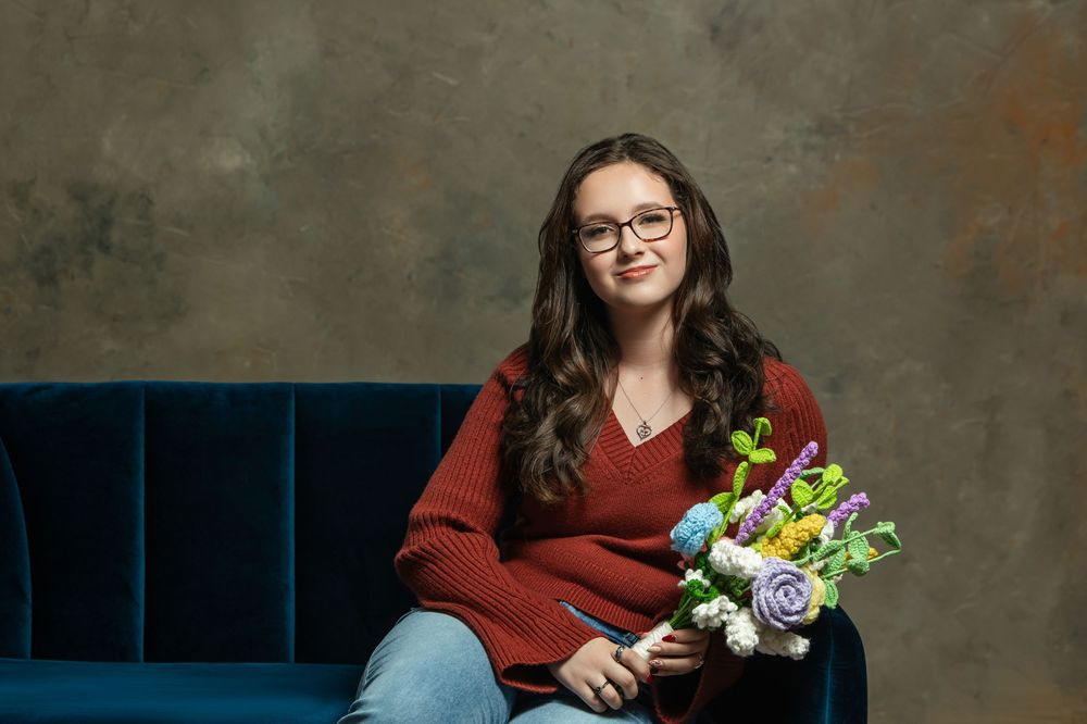 Smiling young woman with glasses holding colorful flowers while seated on a blue velvet sofa.