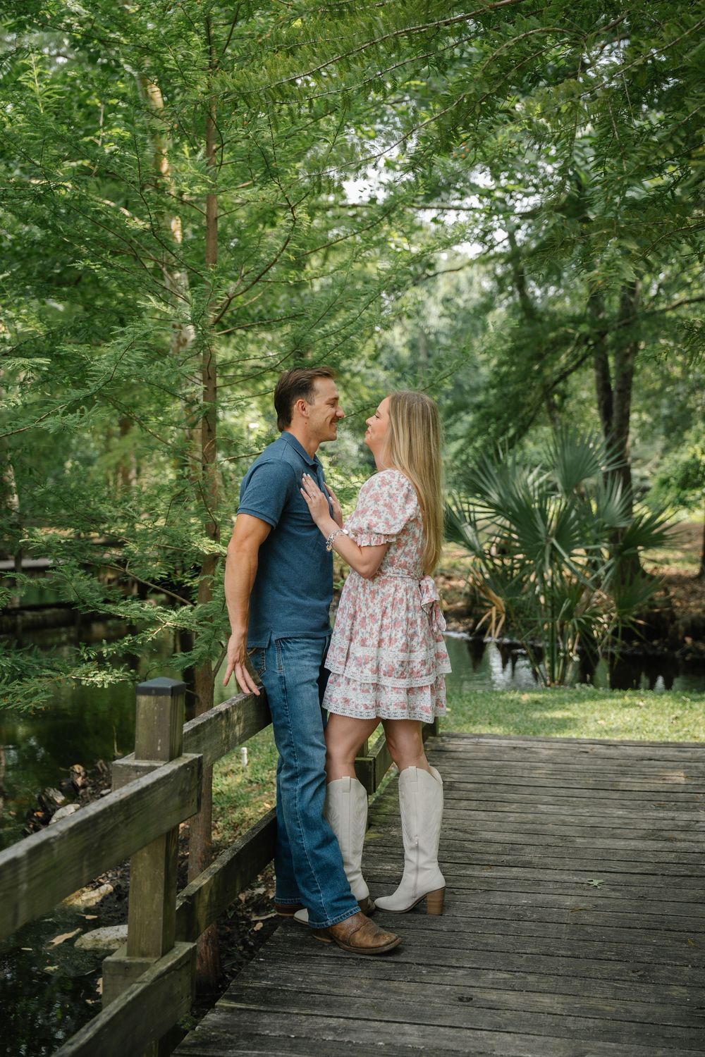 Couple embracing on a wooden bridge surrounded by lush green trees near a pond.