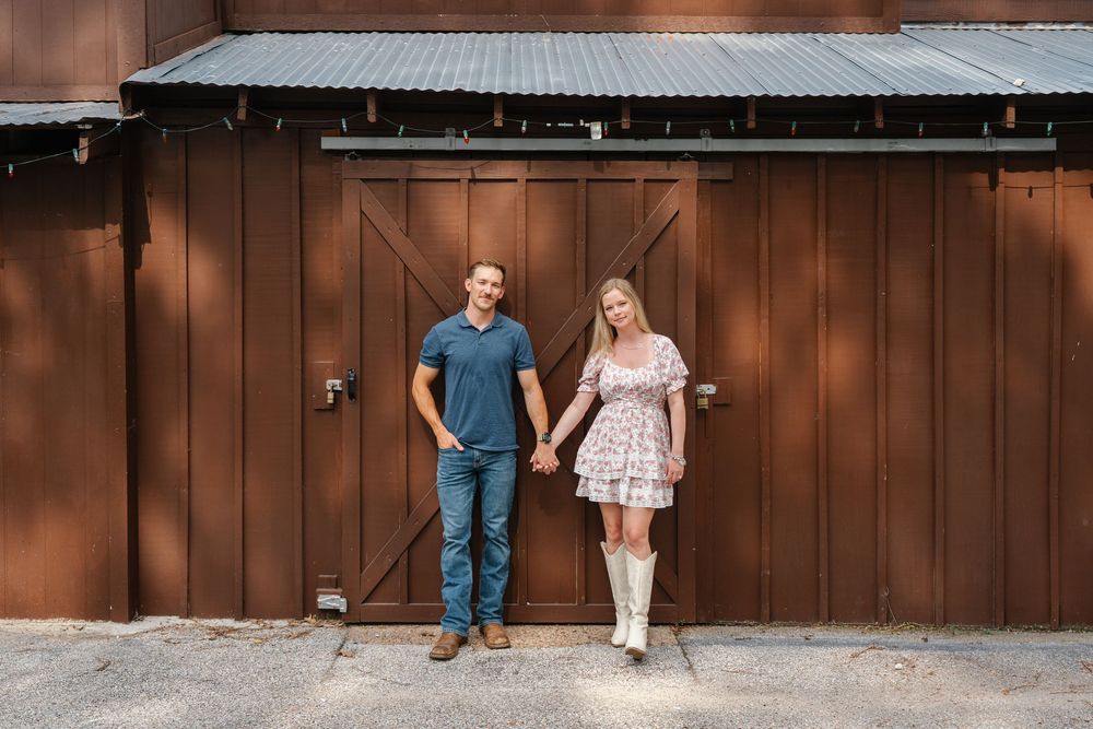 Engaged couple holding hands in front of rustic brown barn doors, woman wearing white cowboy boots.