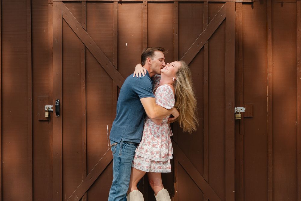 Couple sharing a passionate kiss in front of wooden barn doors during an outdoor engagement session.