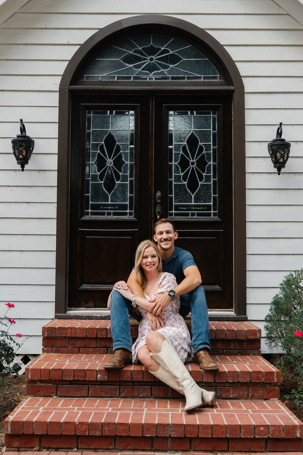 Smiling couple sitting on brick steps in front of elegant arched double doors with stained glass windows.