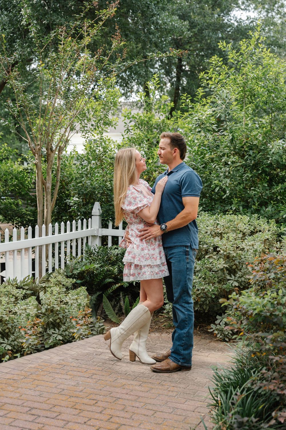 Couple embracing on a brick garden path surrounded by lush greenery and a white picket fence.