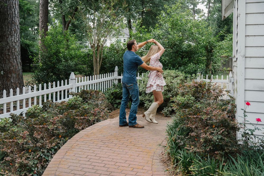 Couple dancing playfully on a brick garden path surrounded by trees and a white picket fence.