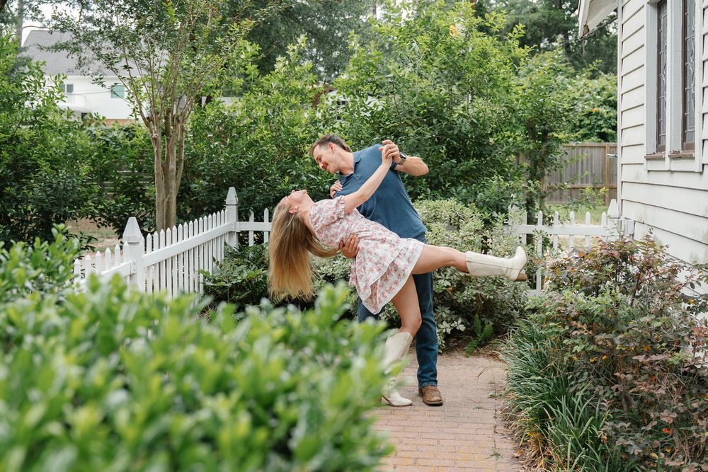 Man lifting and spinning woman in a floral dress on a brick garden path during engagement photos.