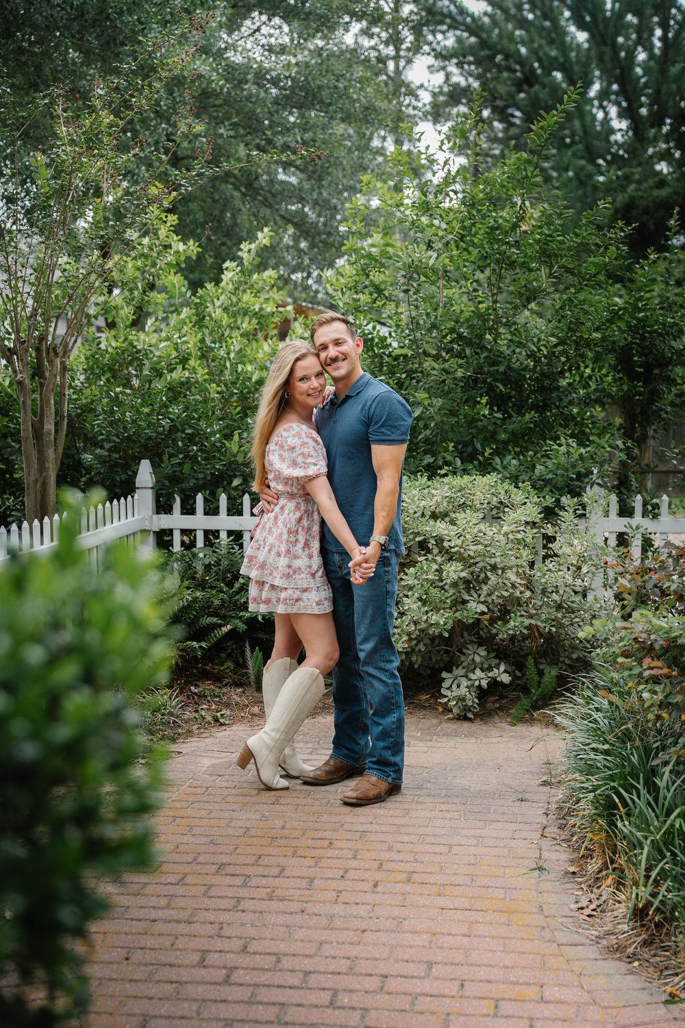 Happy couple holding hands on a brick garden path surrounded by lush green shrubs and white fence.