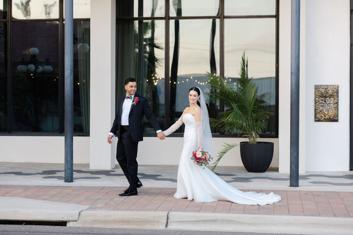 Bride and groom holding hands and walking together outside a modern wedding venue, captured during their wedding day.