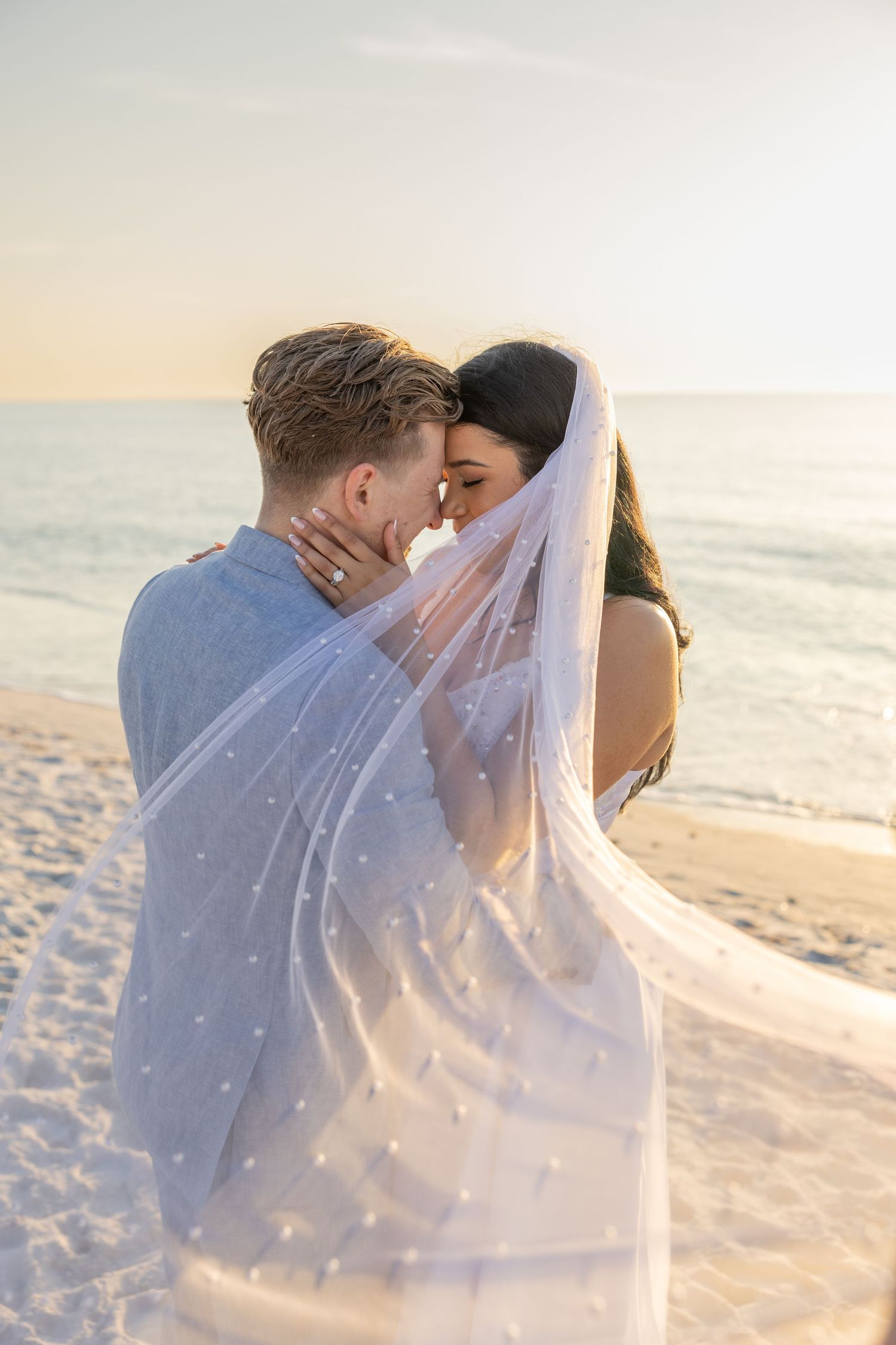 Bride and groom embracing with a veil blowing around them during a beach wedding portrait.