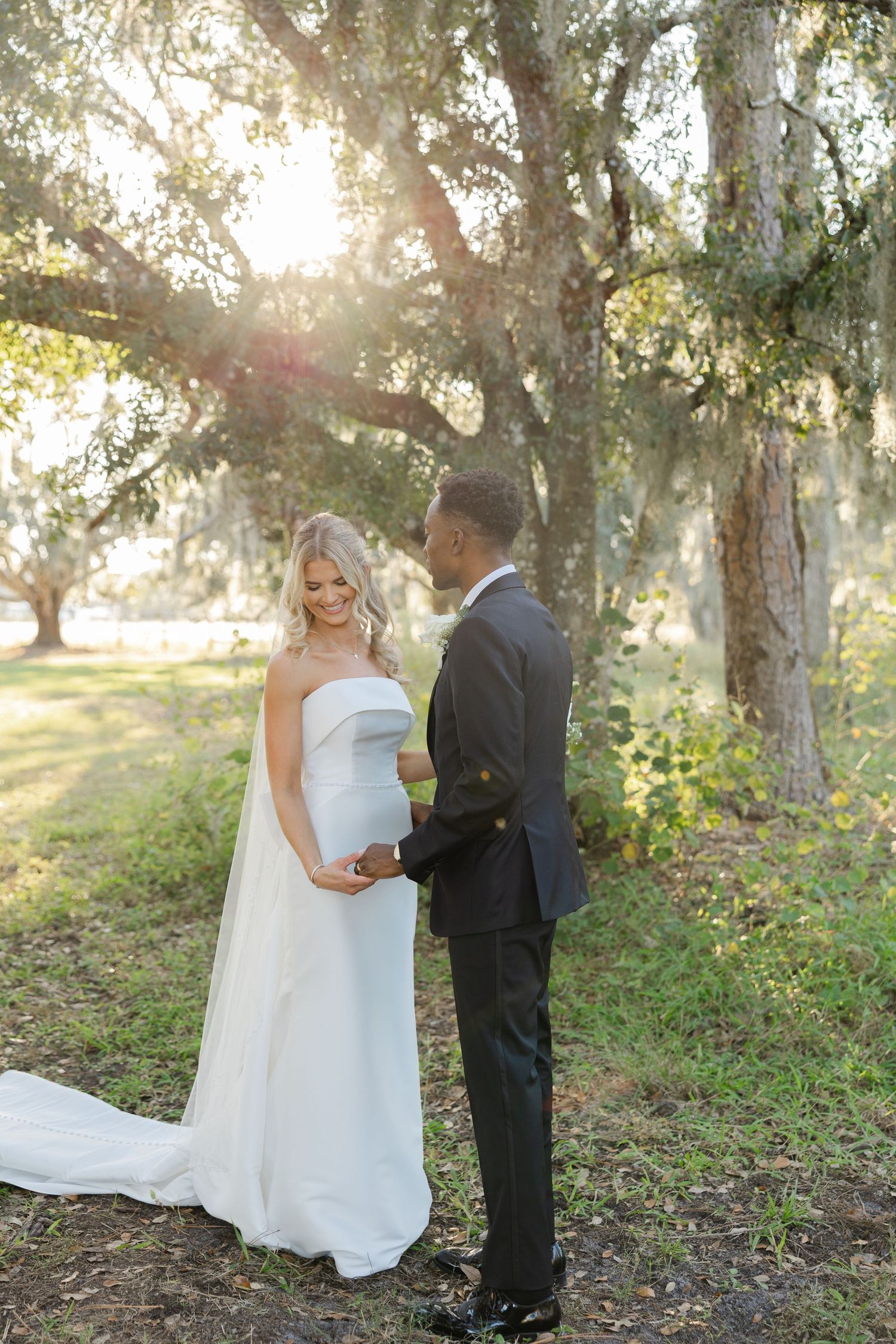 Bride and groom standing together beneath trees with sunlight filtering through during a wedding portrait session.