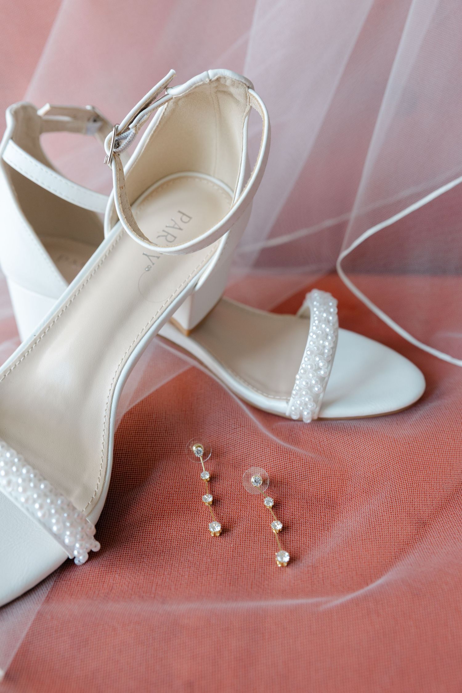 Ivory bridal heels and pearl jewelry styled on a soft pink surface, photographed as elegant wedding day detail imagery.