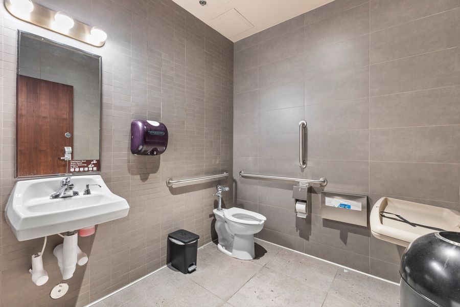 ADA-accessible commercial restroom with grab bars, wall-mounted sink, toilet, and gray tile walls.