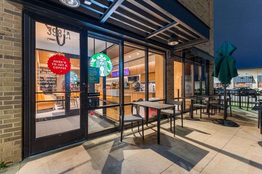 Starbucks coffee shop exterior at night with outdoor patio seating and green umbrellas beside brick storefront.