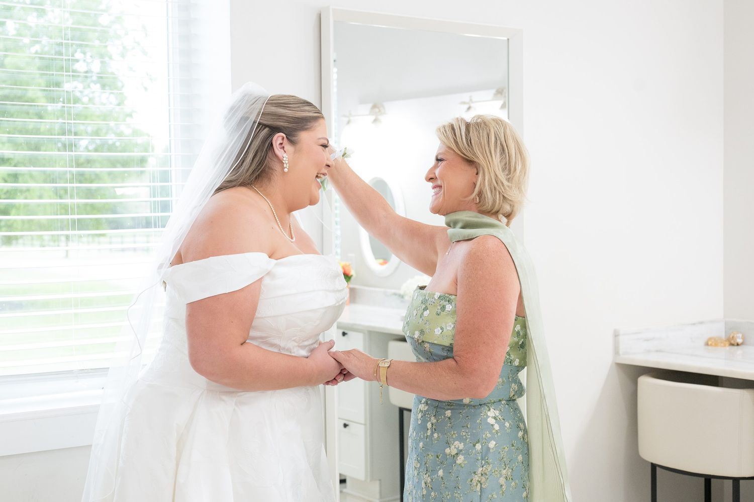 Bride in white gown and veil shares tender moment with mother in floral dress during wedding preparations.