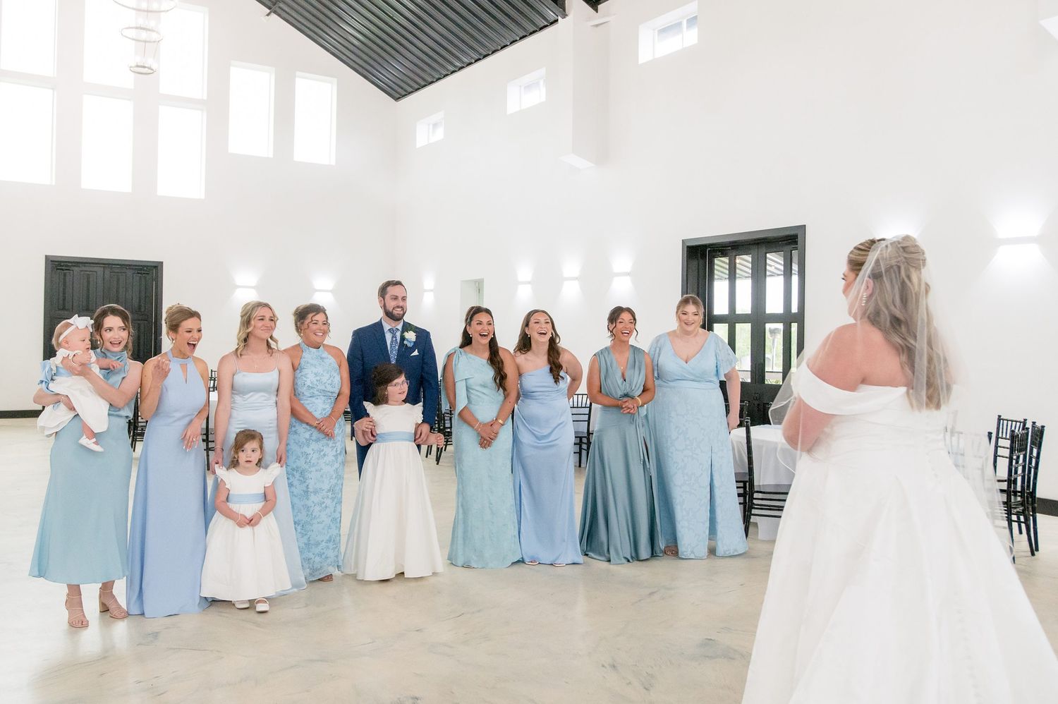 Groom stands with bridesmaids in light blue gowns as bride in white dress makes grand entrance in bright venue.