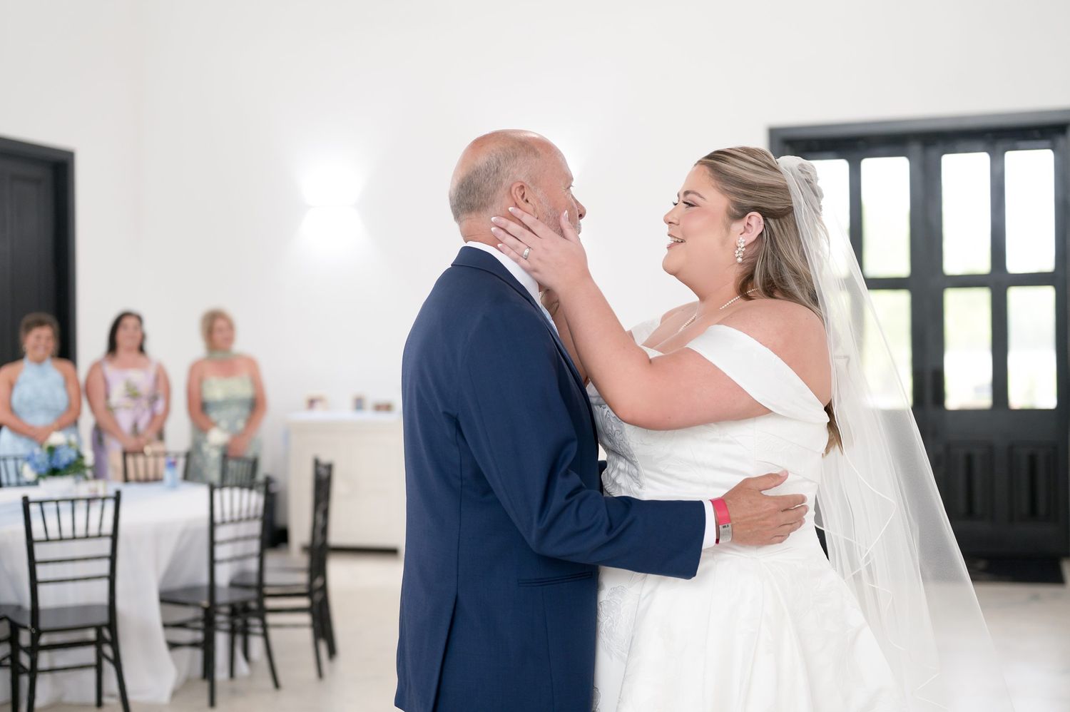 Bride and groom share their first dance, smiling tenderly, with wedding guests seated in background at elegant white venue.
