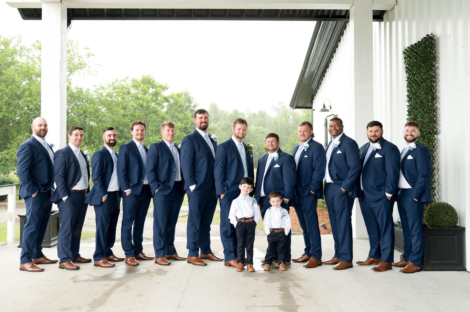 Groom and groomsmen in navy suits with white boutonnieres pose with two young ring bearers outside a white venue.