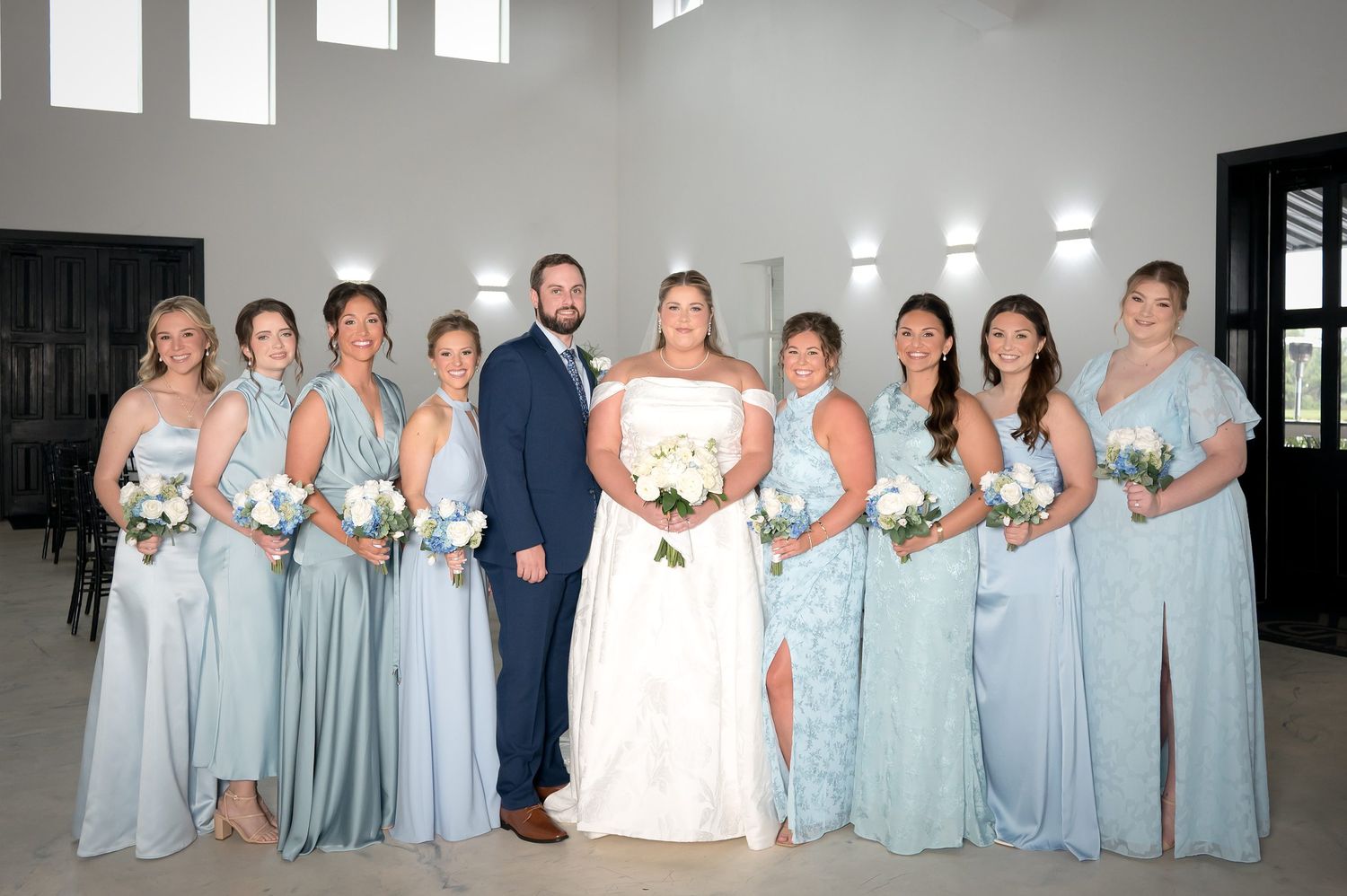 Wedding party poses with bride, groom, and bridesmaids in mismatched light blue gowns holding white bouquets.
