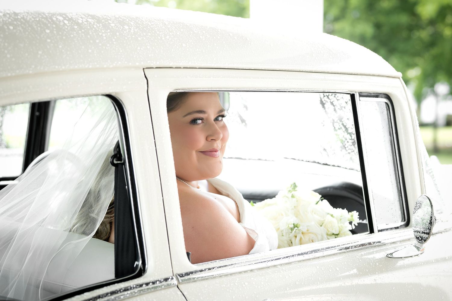 Bride in wedding dress and veil sitting in a vintage white car holding a bouquet.
