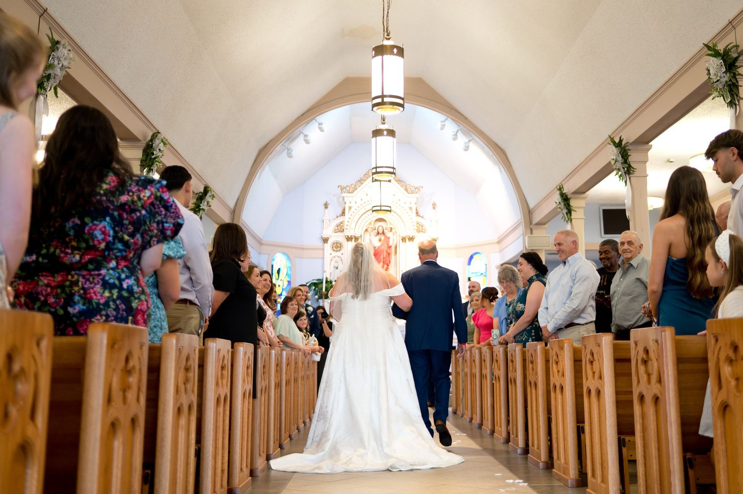 Bride walking down the aisle with escort in a white church wedding ceremony with wooden pews.