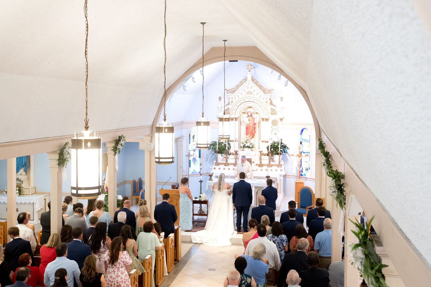 Wide angle view of a wedding ceremony inside a white church with guests seated in pews along the aisle.