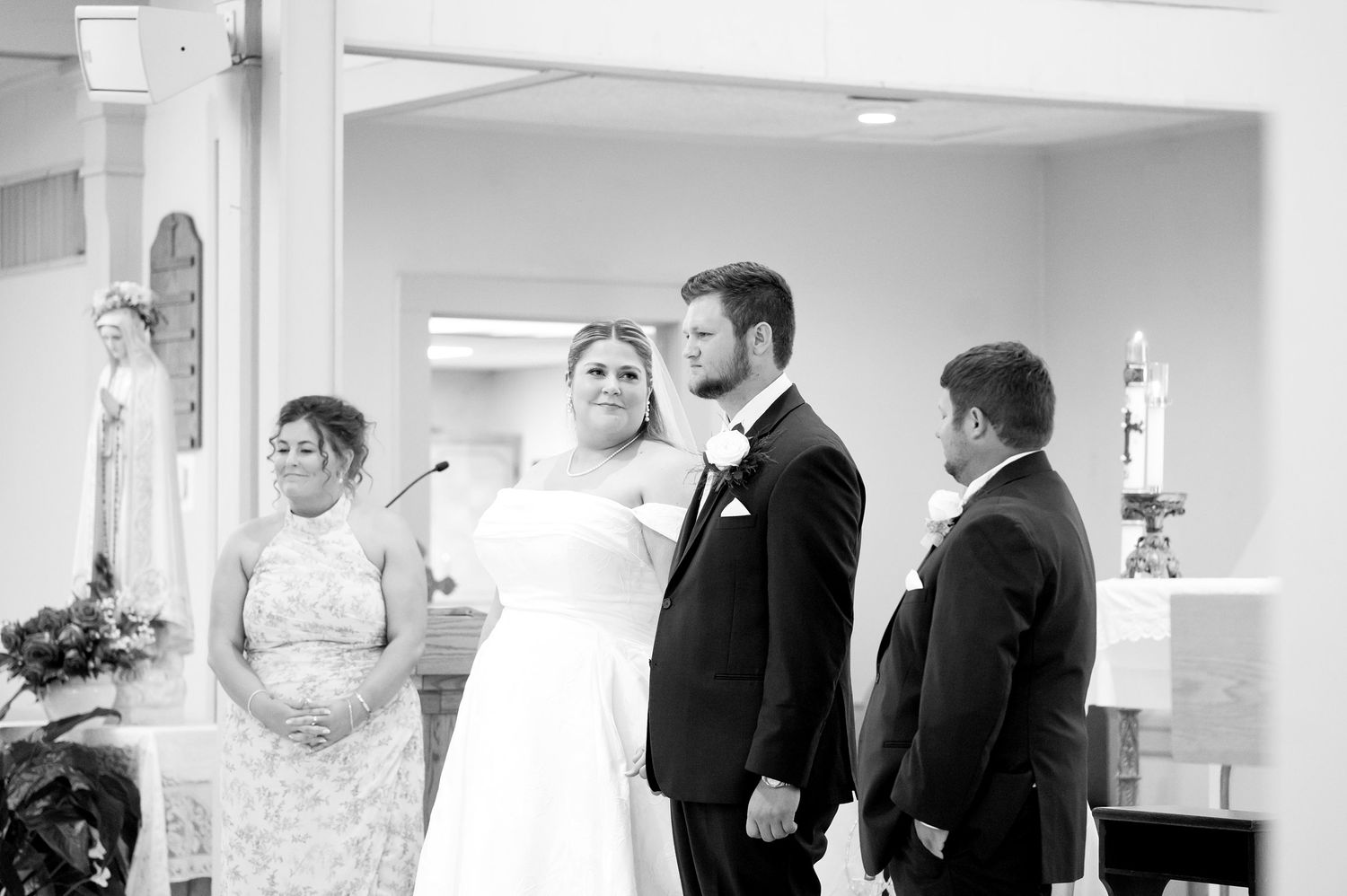 Black and white photo of bride and groom exchanging vows at church altar with officiant and groomsman nearby.