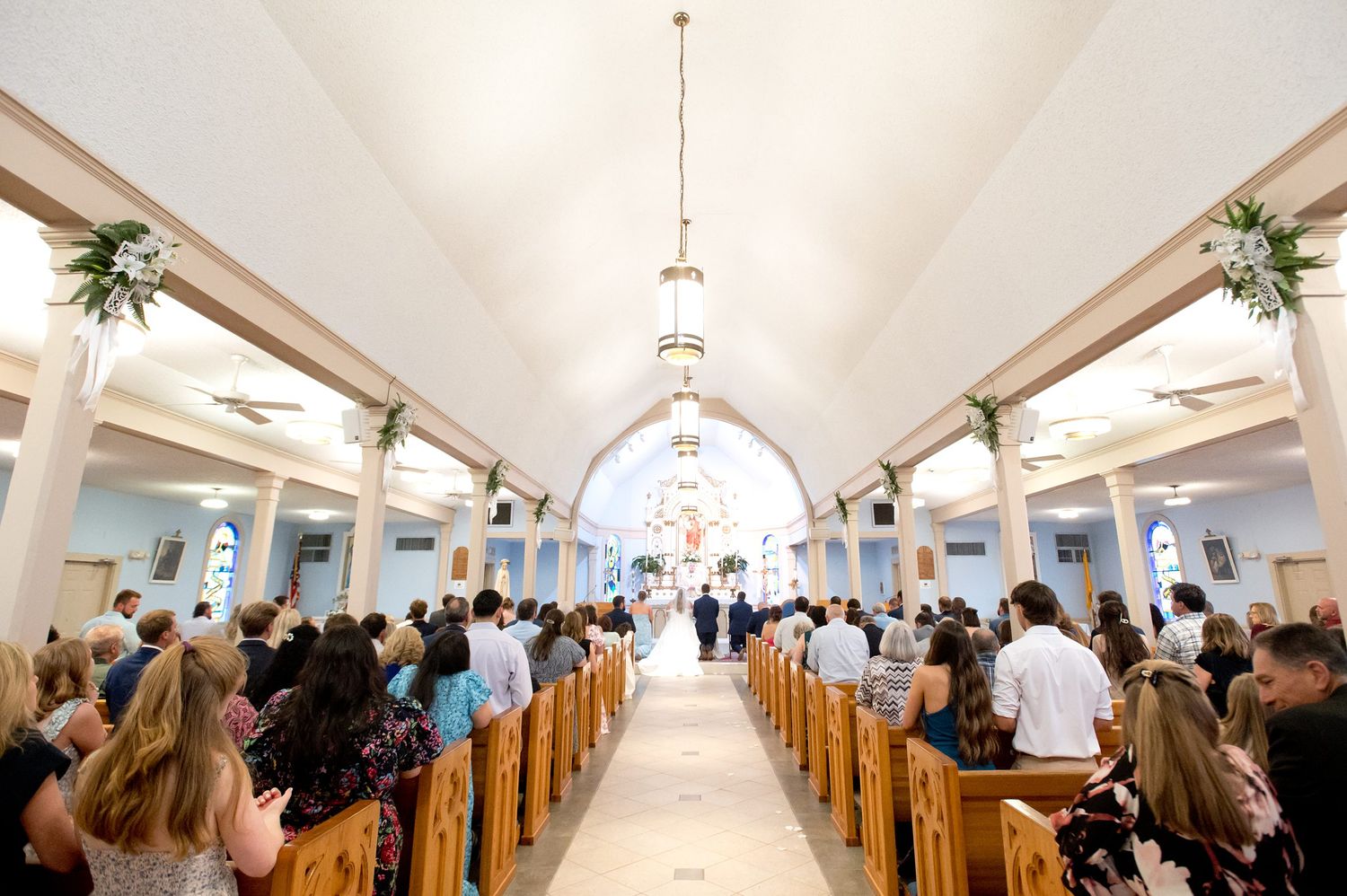 Wide view of a church wedding ceremony with guests seated in wooden pews and couple at the altar.