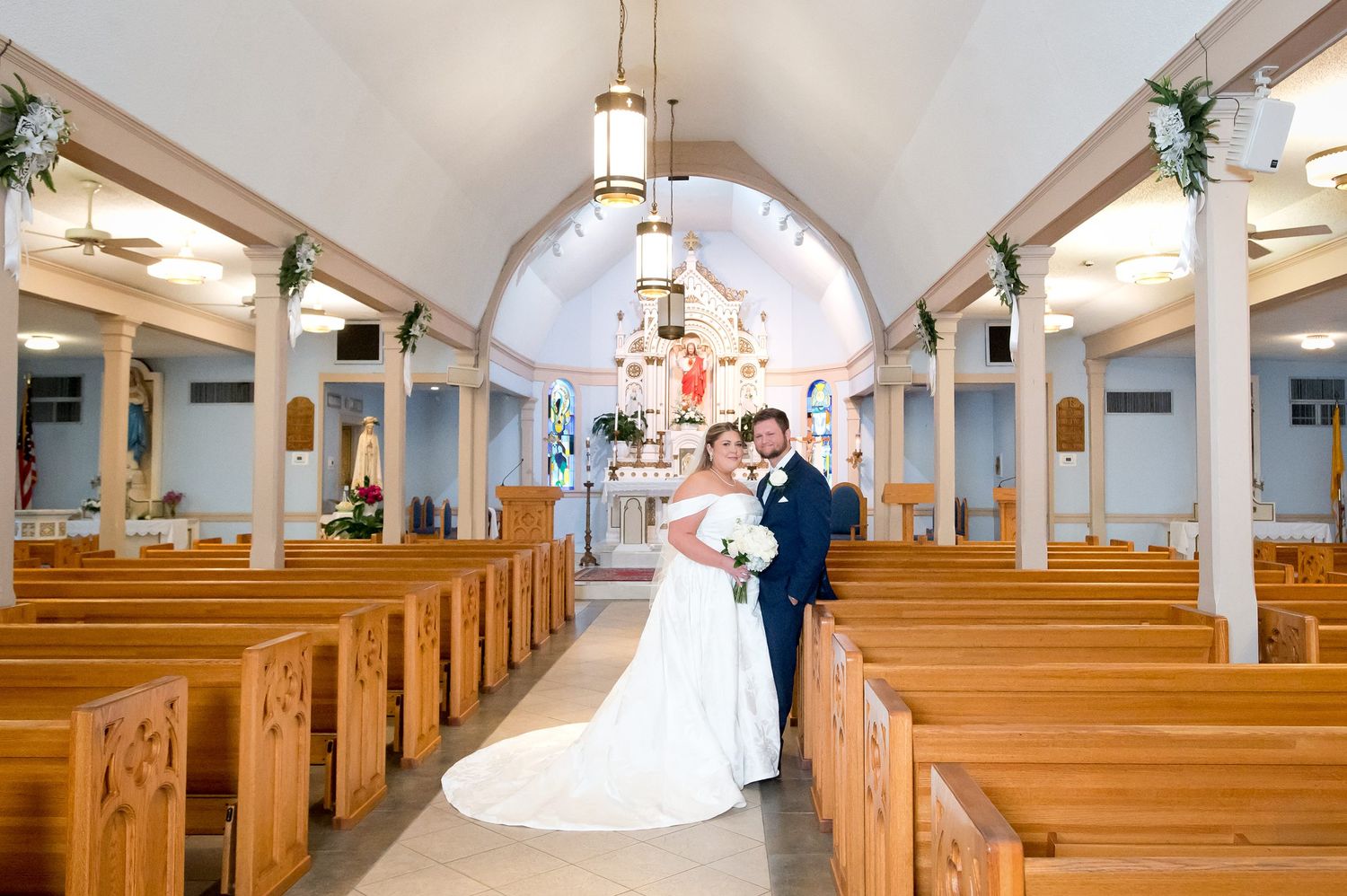 Bride and groom pose together in a sunlit church aisle surrounded by wooden pews on their wedding day.
