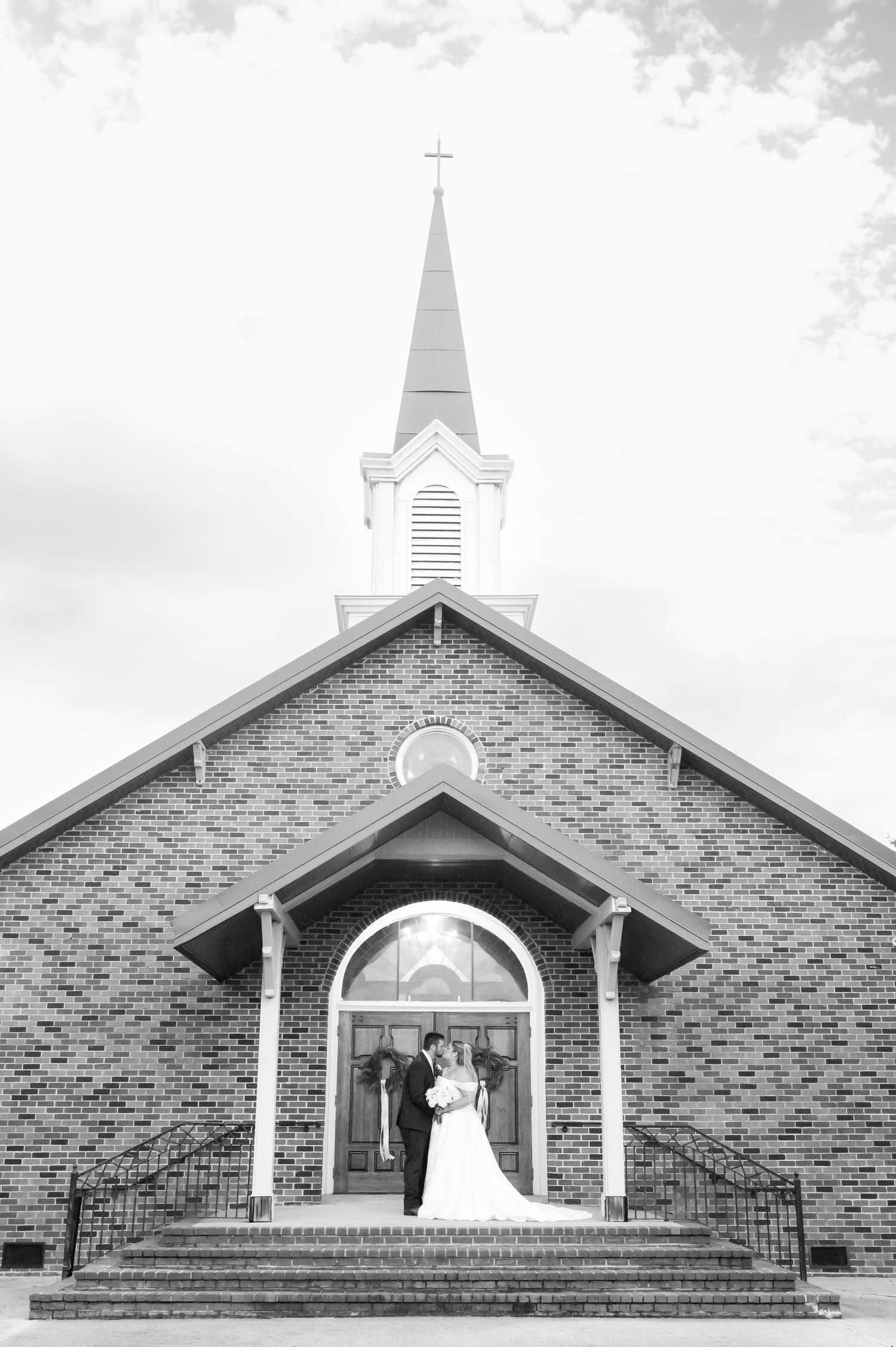 Black and white photo of a bride and groom standing on church steps beneath a brick steeple on their wedding day.