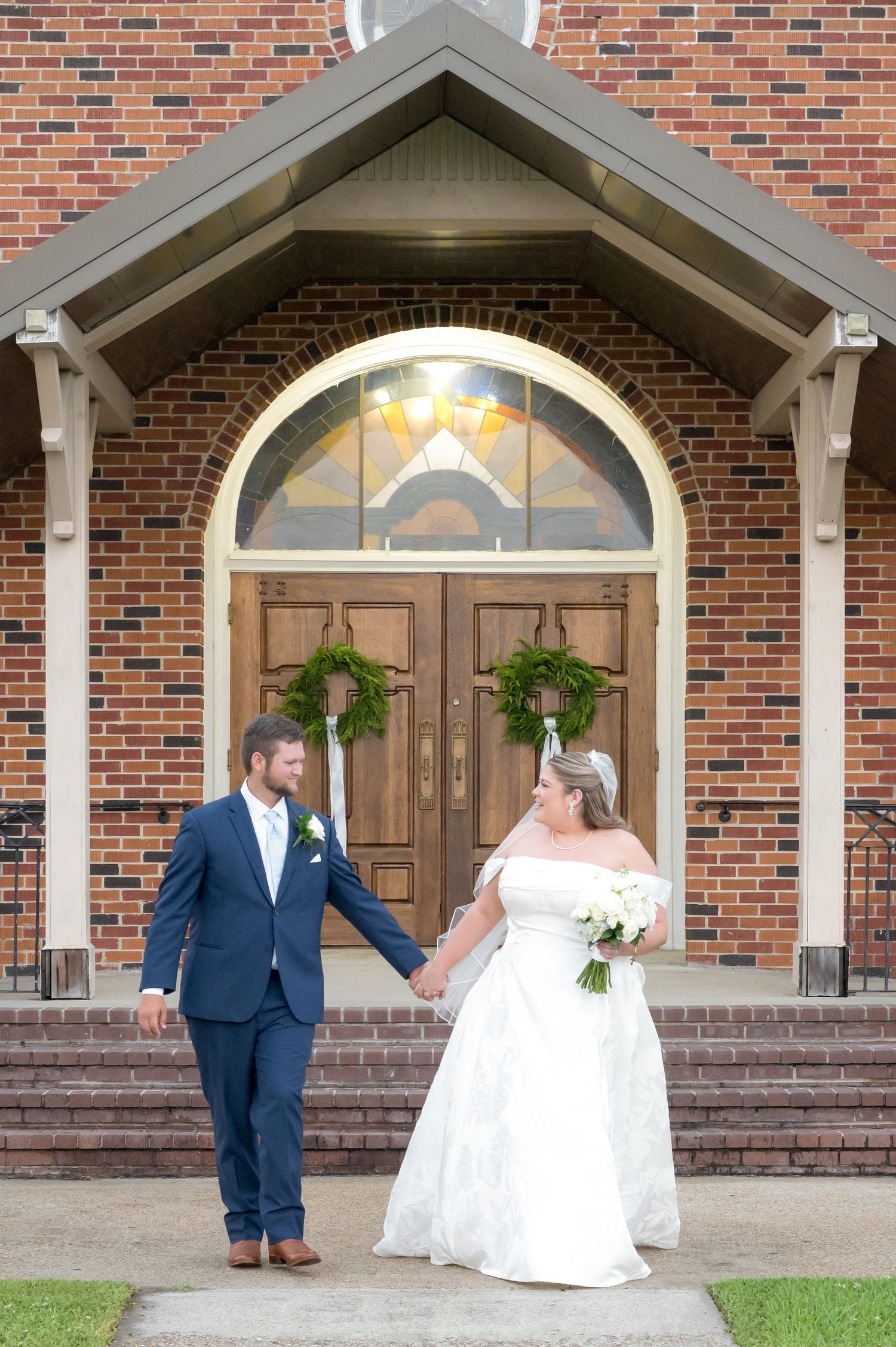 Bride and groom holding hands in front of a brick church with wooden doors and greenery wreaths on their wedding day.