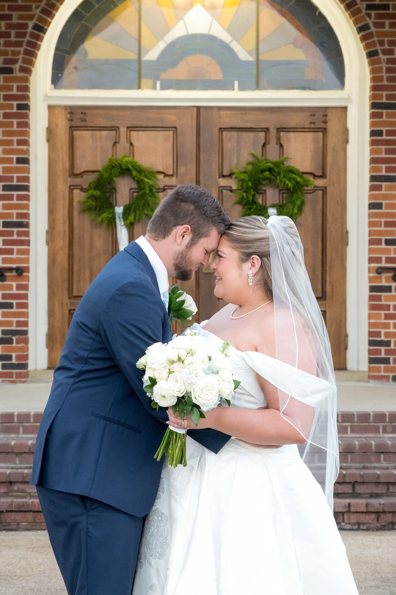 Bride and groom touching foreheads outside brick church doors, bride holding white bouquet and wearing veil.
