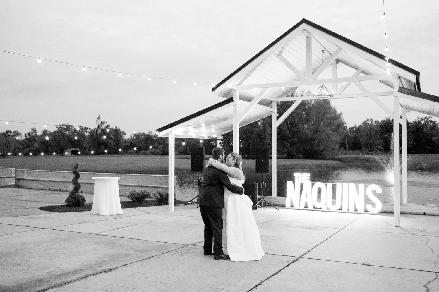 Bride and groom share first dance outdoors at dusk near illuminated Naquins marquee sign at wedding reception.