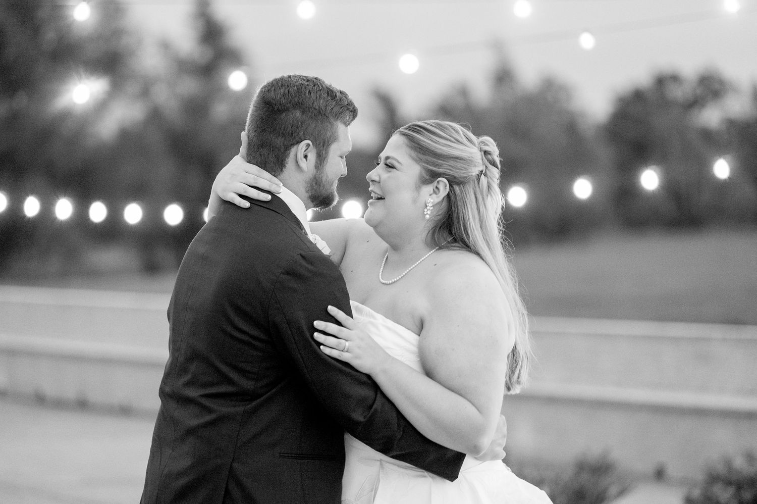 Black and white photo of a bride and groom sharing a romantic dance under string lights at their wedding reception.