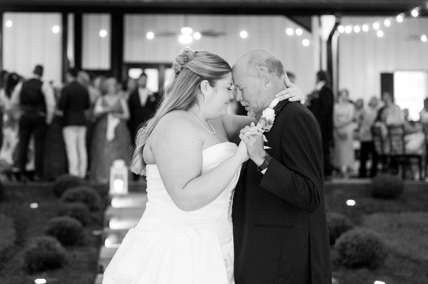 Bride shares emotional father-daughter dance at elegant outdoor wedding reception with string lights glowing.
