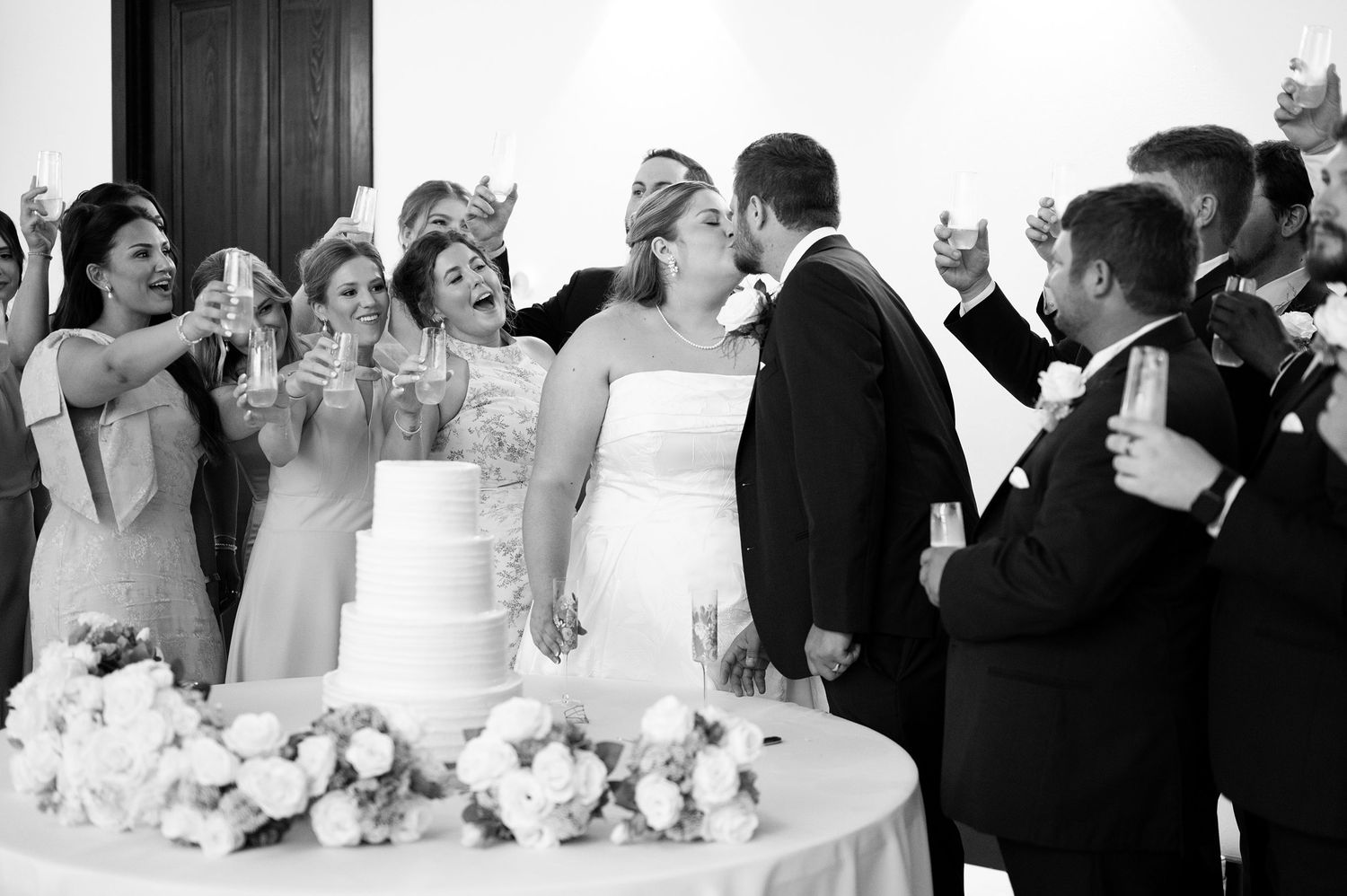 Black and white photo of bride and groom kissing at wedding cake cutting while bridal party celebrates with champagne.