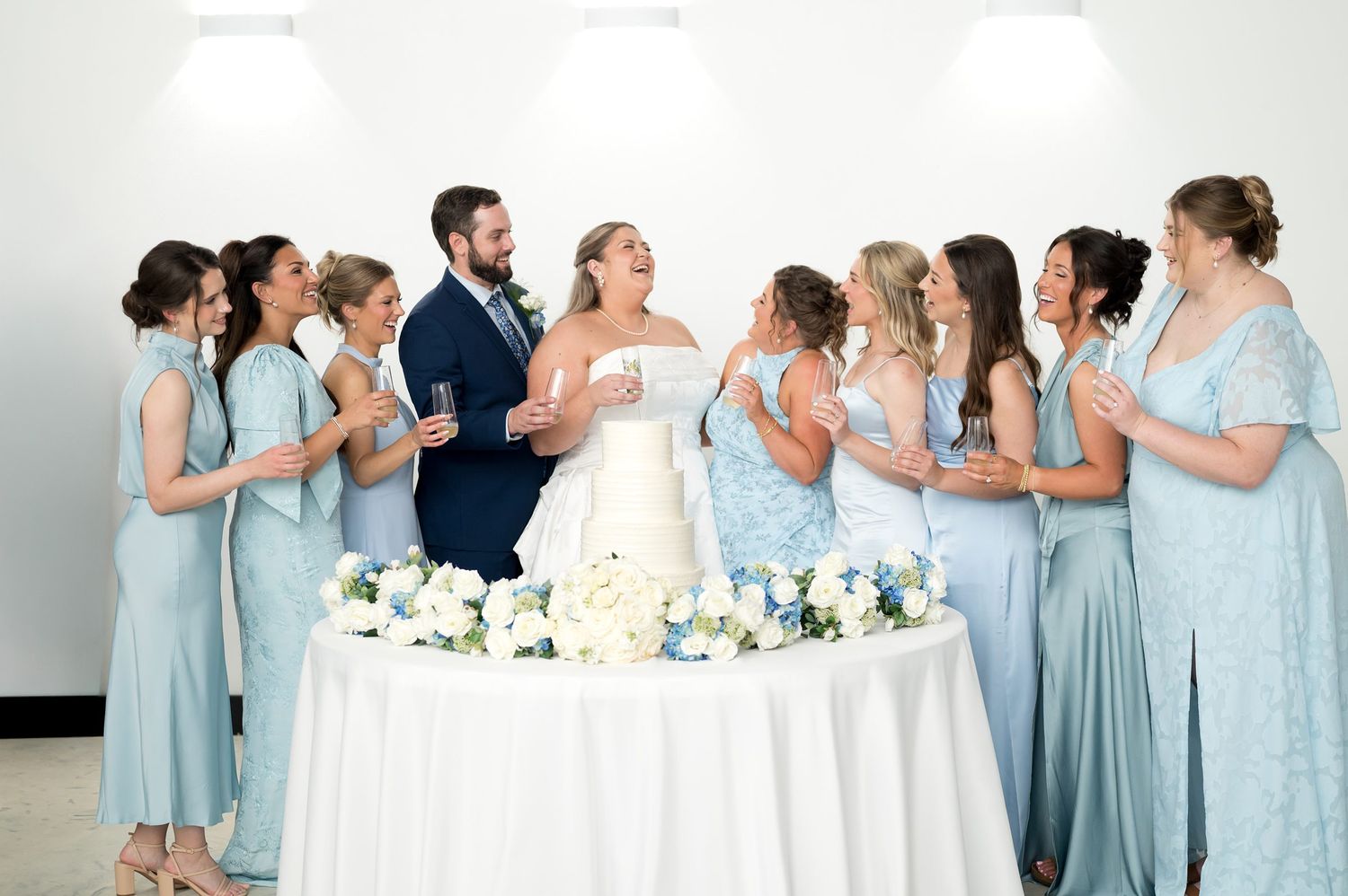 Bride and groom cutting their wedding cake surrounded by bridesmaids in light blue dresses at reception.