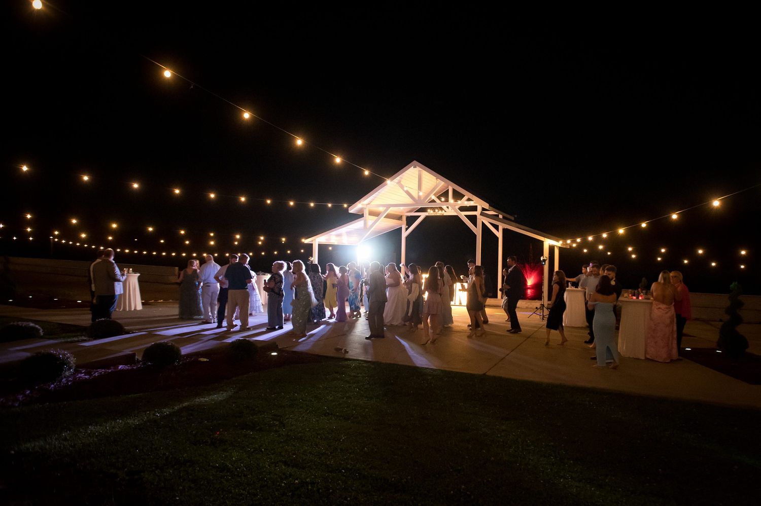 Outdoor evening wedding reception under string lights and a wooden pavilion with guests gathered on a lawn at night.