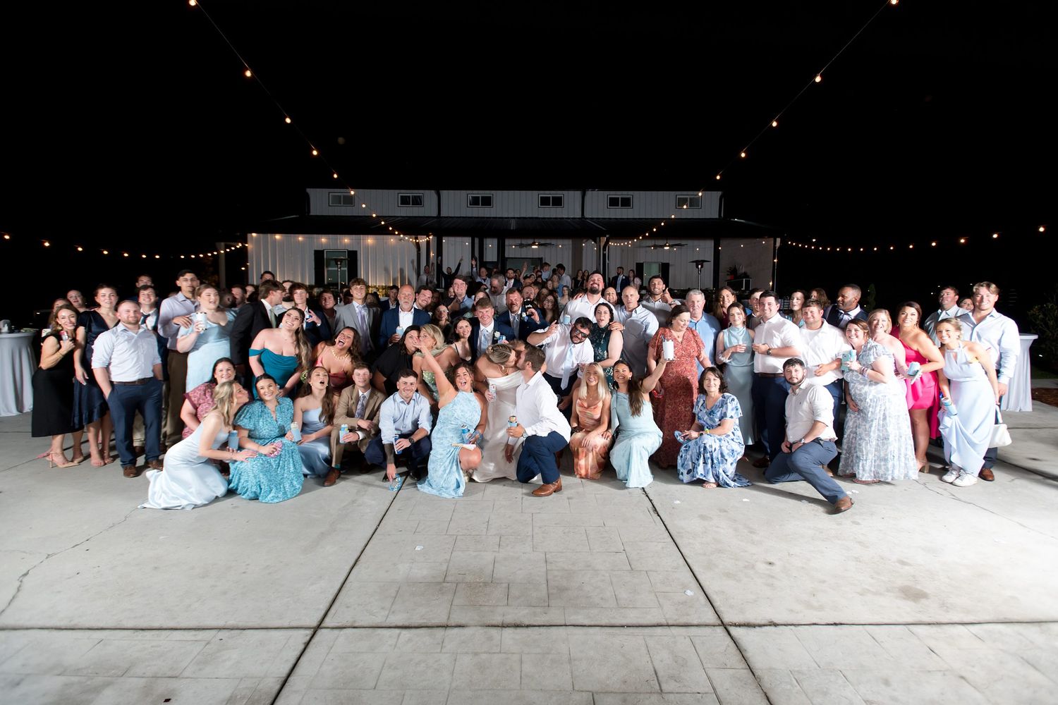 Large wedding group photo outdoors at night with string lights, guests in formal attire posing together.