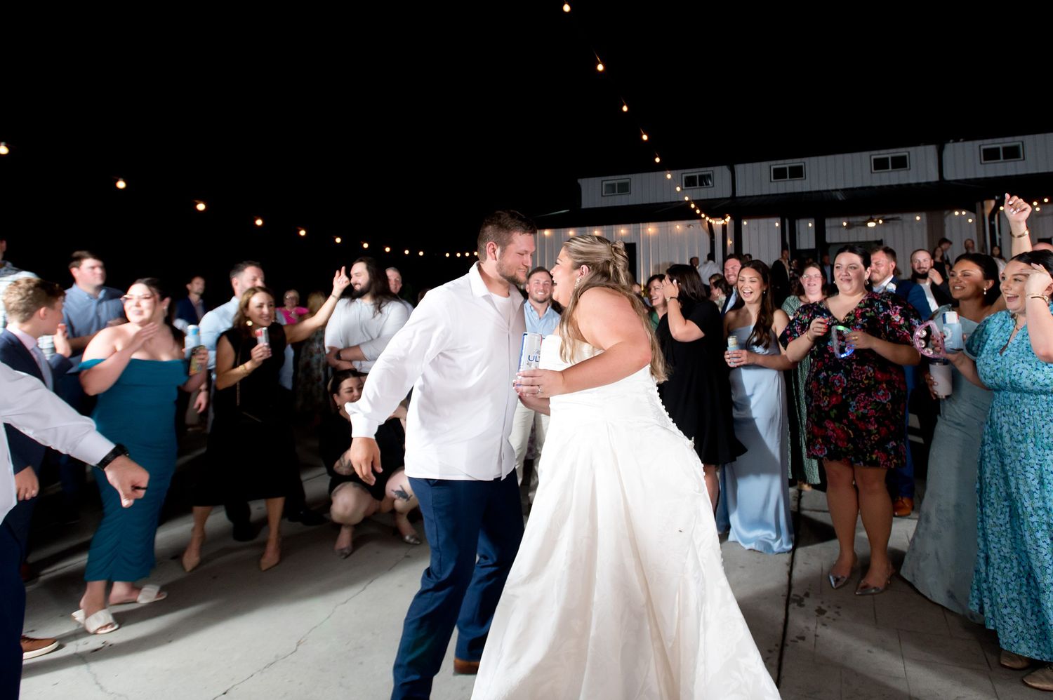 Bride and groom share first dance at outdoor wedding reception under string lights with guests celebrating.