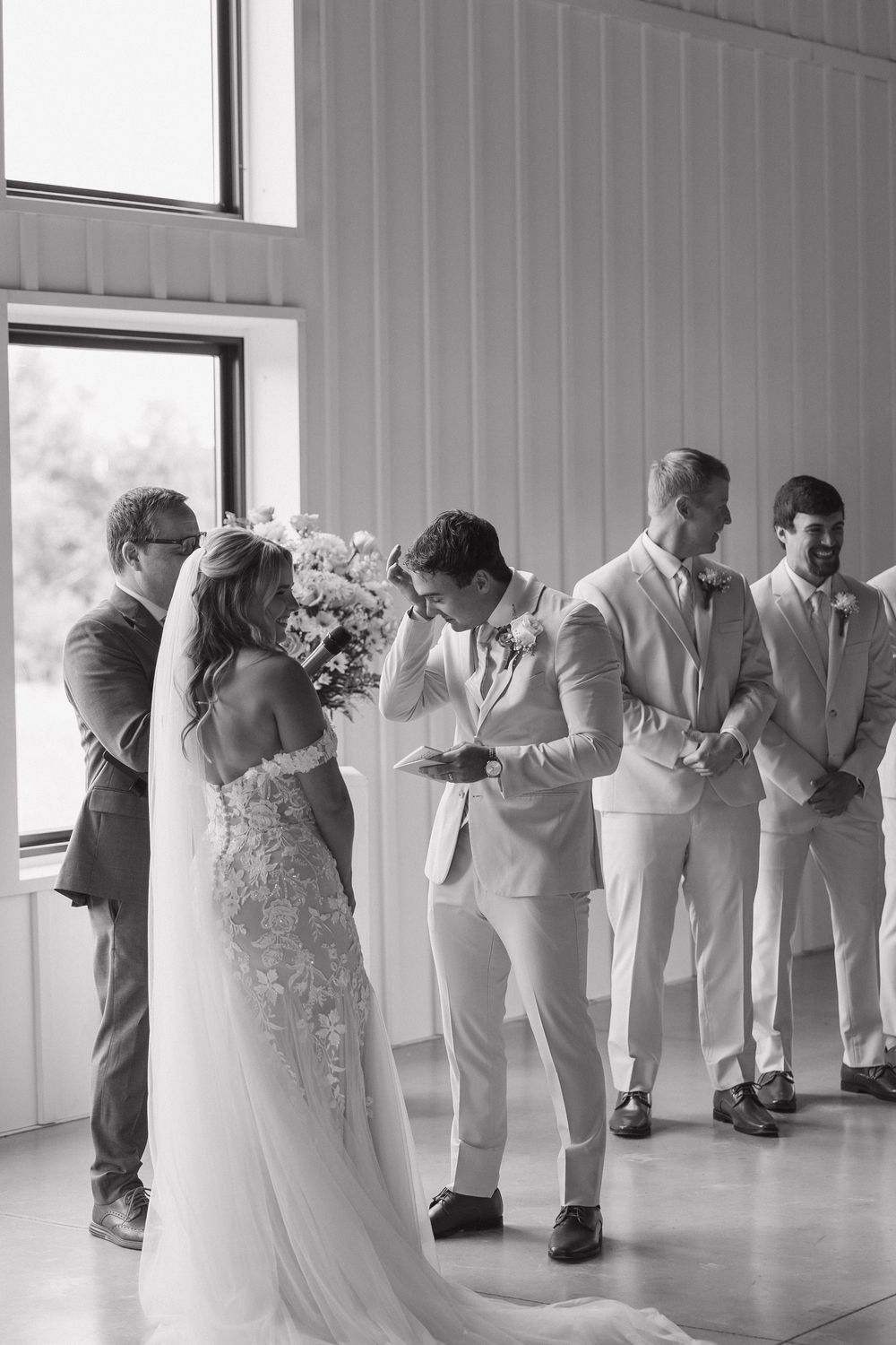 Black and white photo of a groom wiping tears during wedding ceremony with bride and groomsmen standing nearby.
