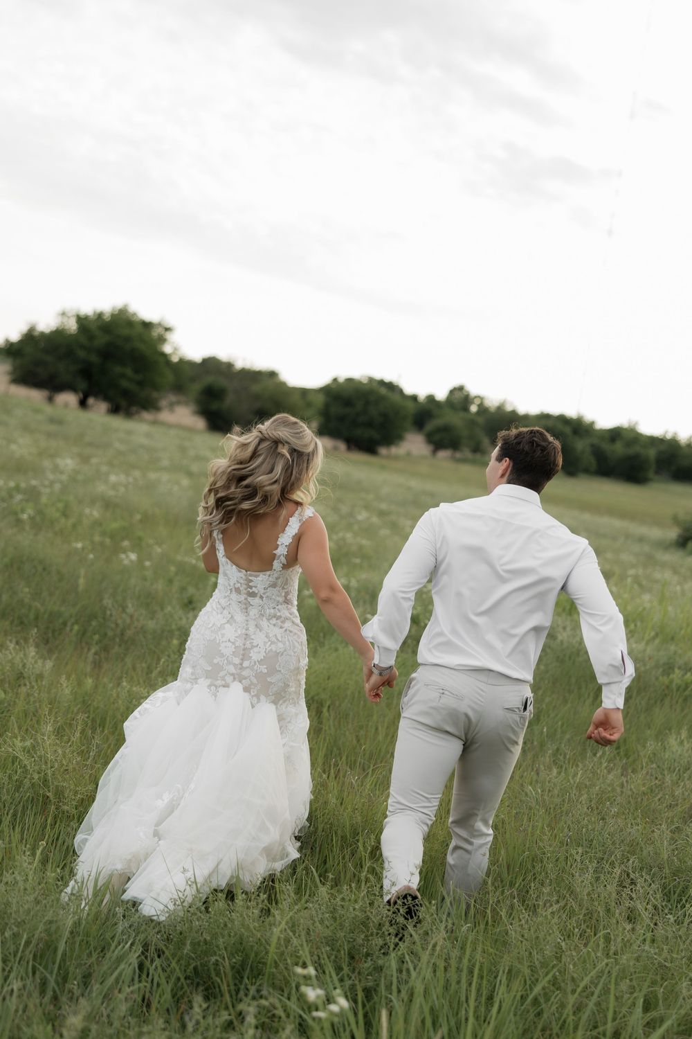Bride and groom holding hands walking through a lush green meadow, bride in lace mermaid gown, groom in white shirt.
