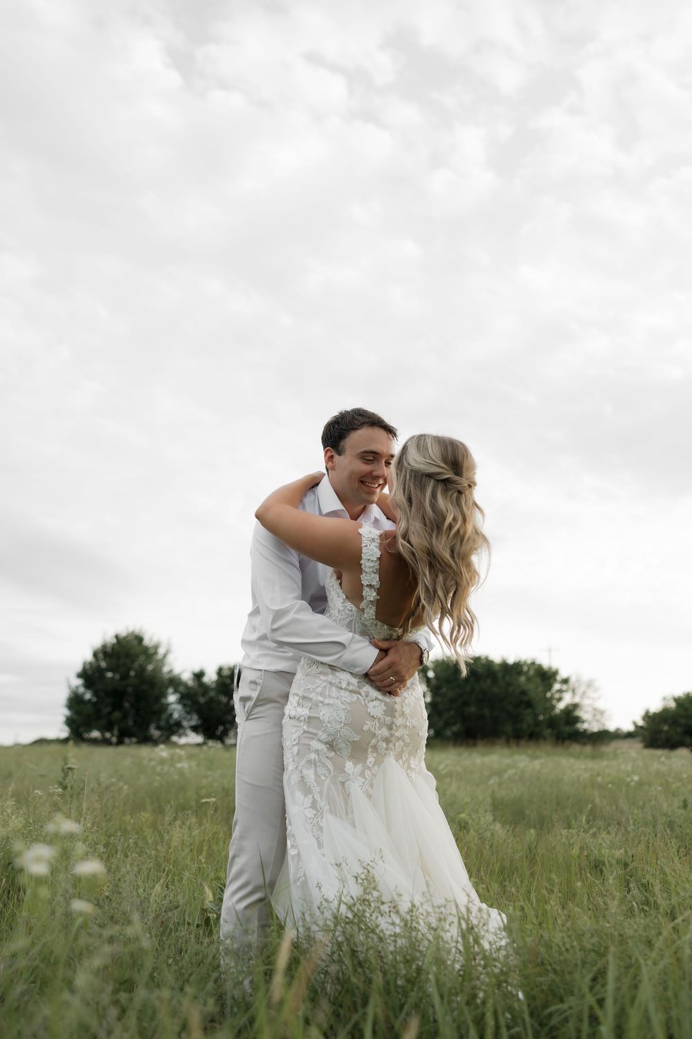 A couple embraces in a lush green field, the bride in a lace gown and groom in gray, sharing a joyful wedding moment.