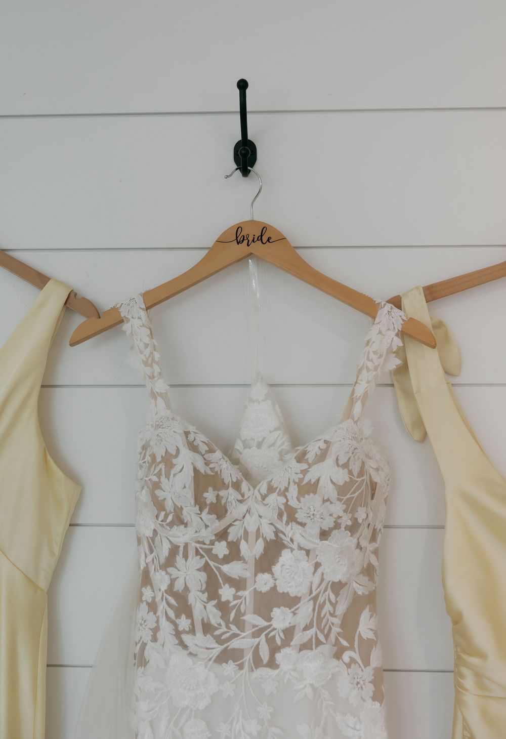 Wedding dress with floral lace detail hangs on wooden bride hanger beside yellow bridesmaid dresses on wall.