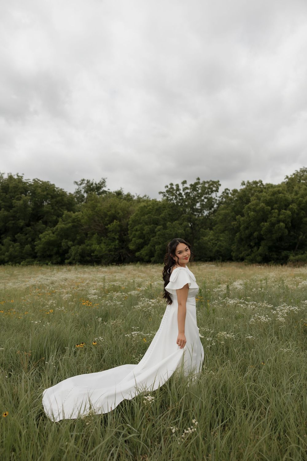 Bride in elegant white flutter-sleeve gown with train standing in wildflower meadow under cloudy sky.