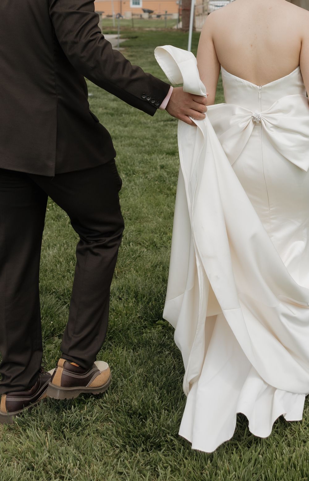 A groom in a dark suit helping carry the train of a bride's white bow-back wedding gown.