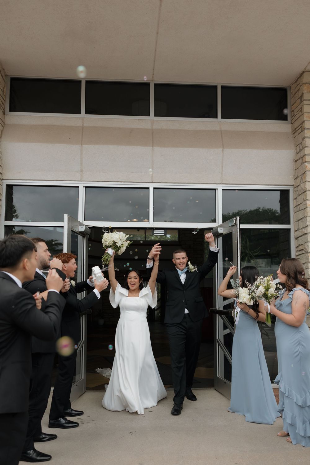 Joyful newlyweds raising hands as they exit church doors surrounded by cheering wedding party members.