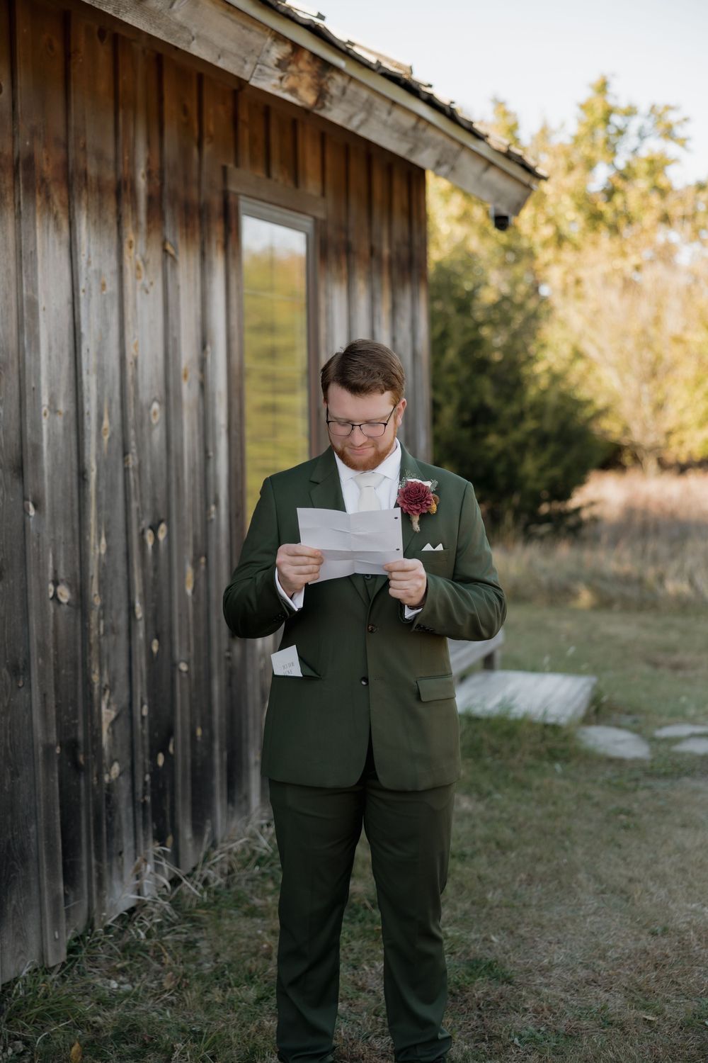 Groom in a green suit reads heartfelt wedding vows from a letter outside a weathered wooden barn.