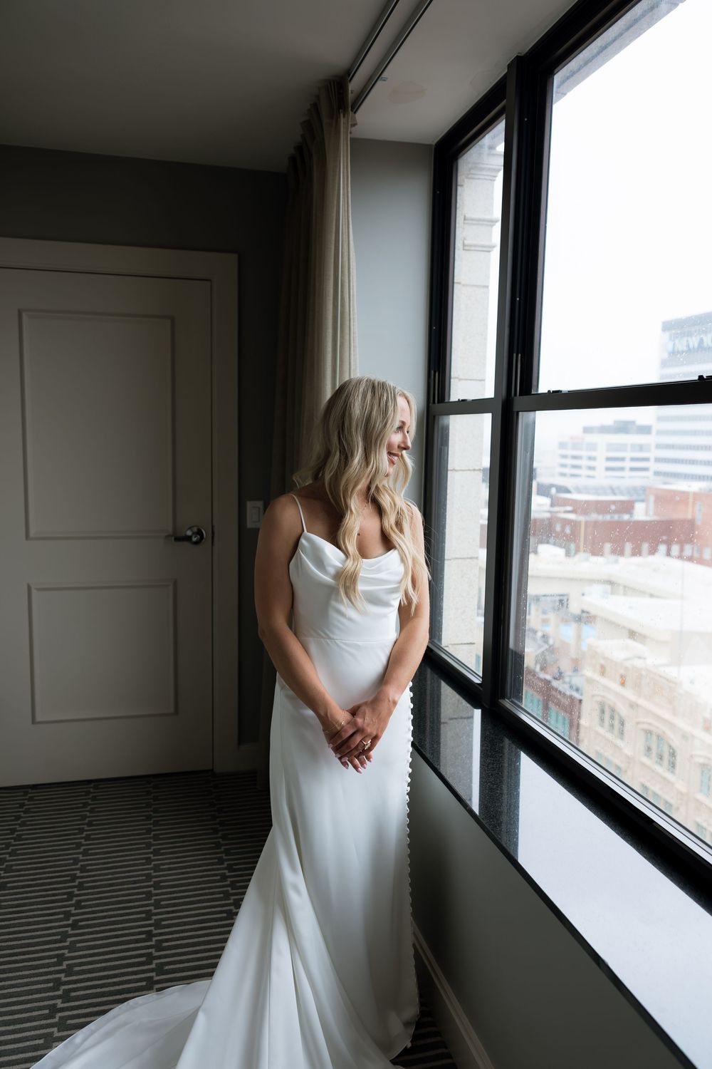 Elegant bride in white satin gown gazing out large hotel room windows.